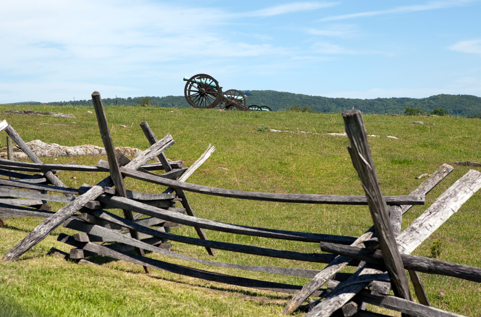 Historic Civil War battlefield at Antietam with split rail fence in the foreground and cannons positioned on a grassy hill under a blue sky