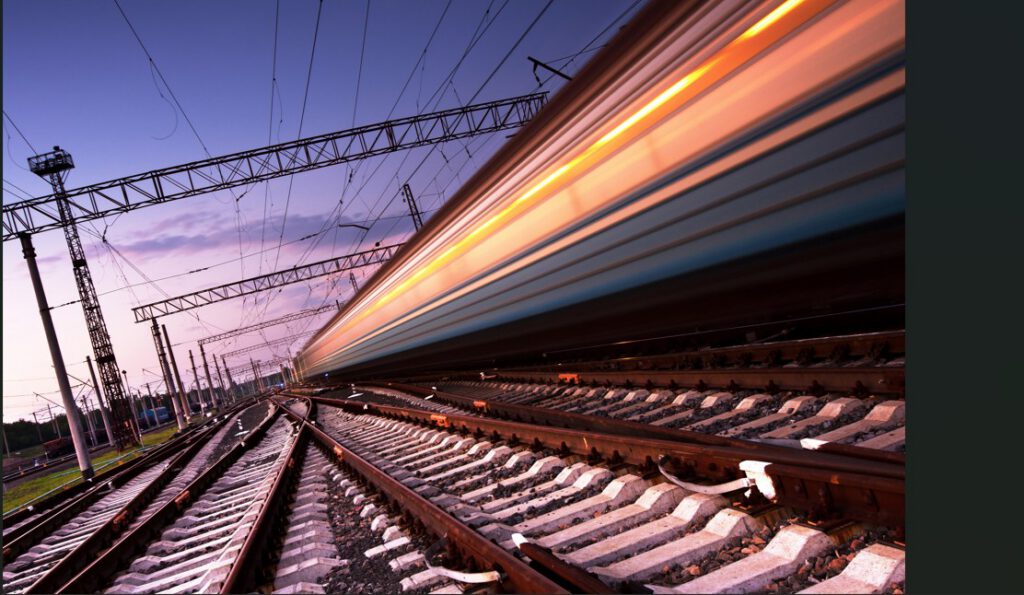 A high-speed train in motion at dusk, captured with a motion blur effect over intersecting railway tracks and overhead electric lines.