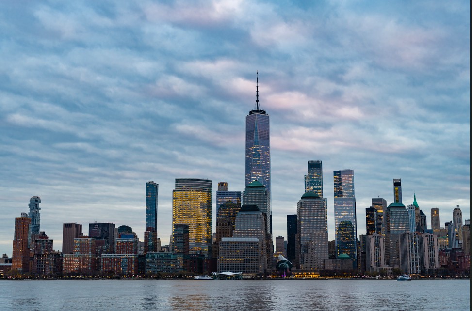 View of the Lower Manhattan skyline at sunset with the One World Trade Center prominently in the center and reflections on the Hudson River.