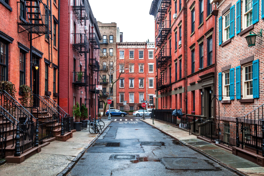 Quaint residential street in Greenwich Village, New York City, lined with red-brick townhouses, iron staircases, and fire escapes on a rainy day