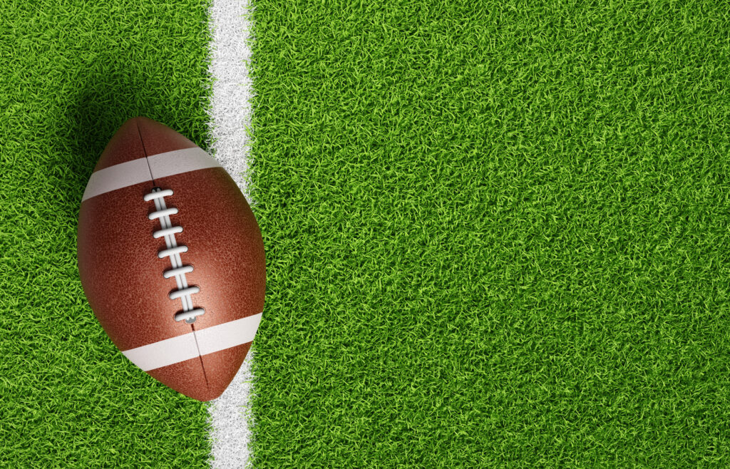 An American football resting on a green football field with yard lines visible