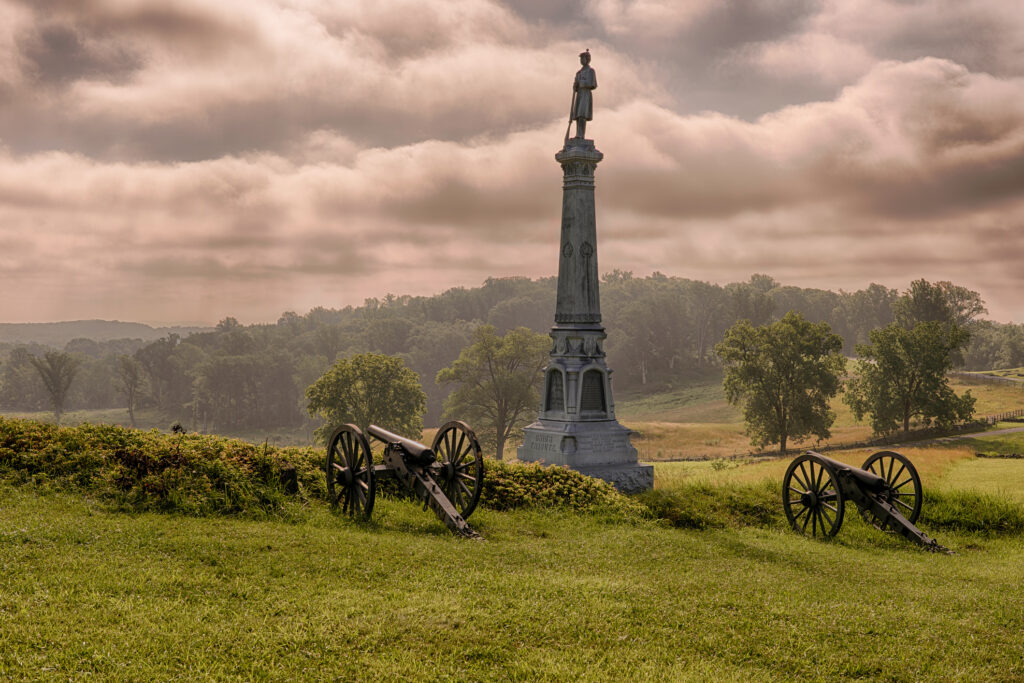 Gettysburg battlefield scene at sunrise with cannons flanking a Civil War monument