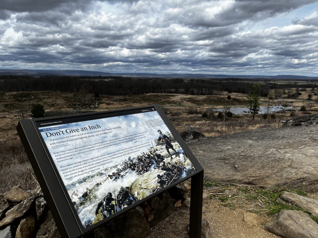 Interpretive sign titled 'Don't Give an Inch' at Little Round Top in Gettysburg, overlooking the historic battlefield under a dramatic cloudy sky