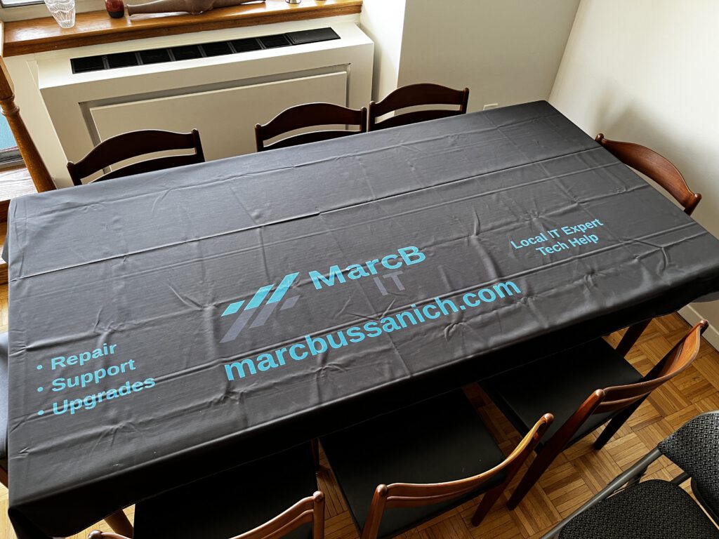 Black tablecloth with MarcB IT logo and website, displayed on dining table before community event setup