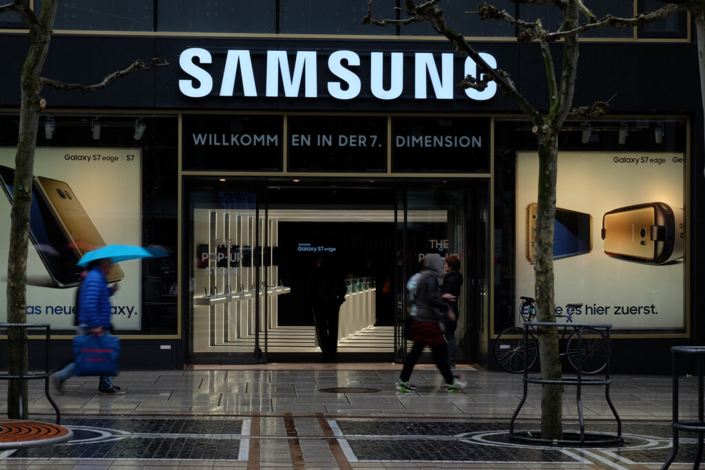 Pedestrians walking past a Samsung storefront in Germany on a rainy day, with a pop-up store entrance and Galaxy S7 Edge advertisements displayed in the windows