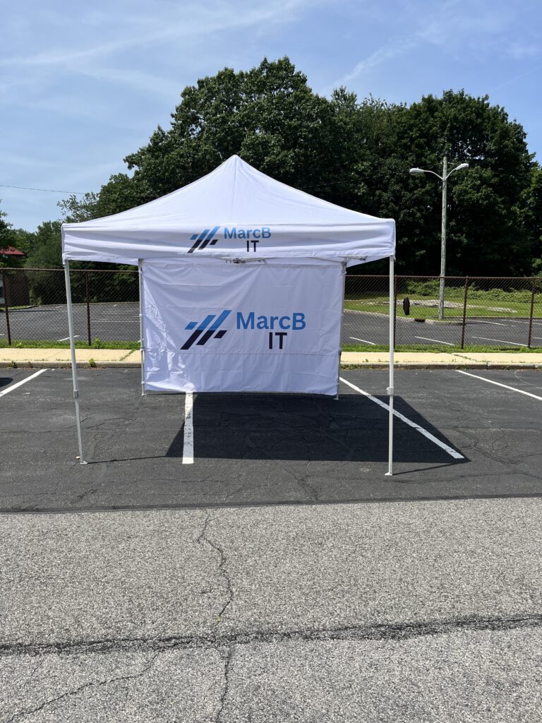 MarcB IT branded white canopy tent set up in a parking lot, featuring the company logo on both the canopy top and back wall, in preparation for the Nyack Street Fair